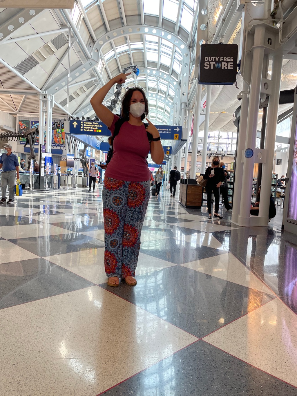 Sarah, a brunette in exercise pants, stands in the terminal of an airport, pointing at decorative dinosaur bones of a brontosaurus, who also wears a mask.
